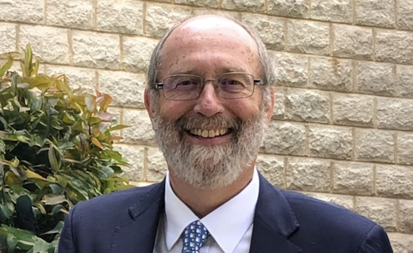 Headshot of Ken Hazel - a smiling white man in glasses with a grey beard, wearing a blue suit and tie.