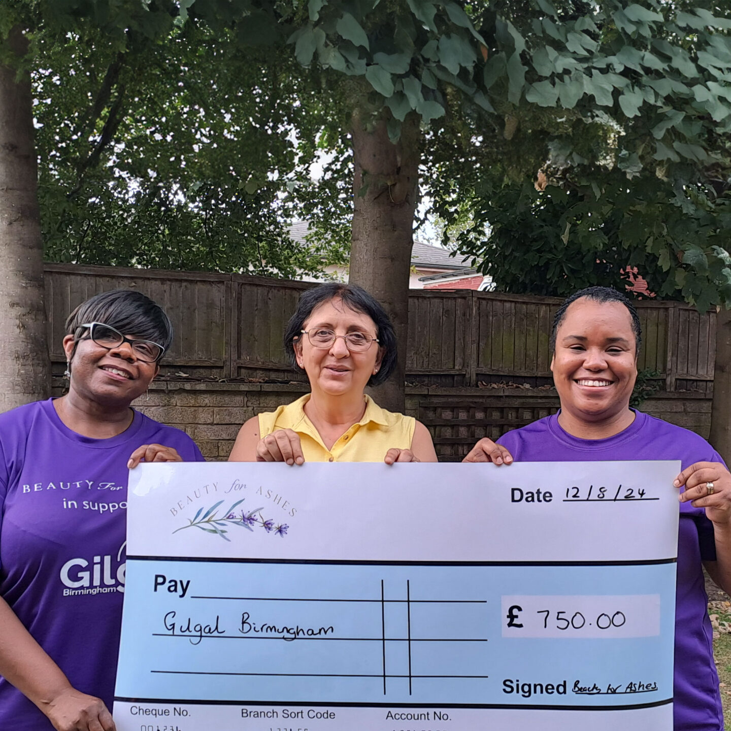 Three women stand outdoors holding a large check made out to "Gilgal Birmingham" for £750. The lady in the middle is Sanja, Gilgal's previous CEO, and the other two women are from Beauty for Ashes.