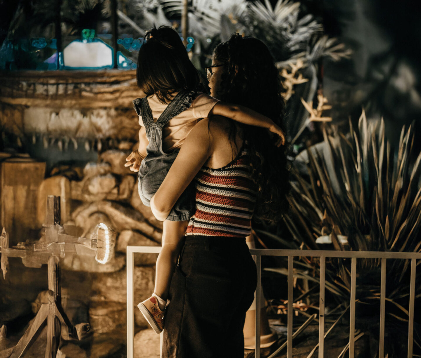 A woman holds a young girl in her arms, both looking at a colorful display in a dimly lit environment with plants in the background.