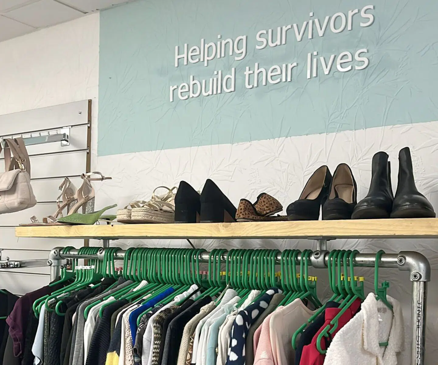 A display in the Gilgal Charity Shop featuring various shoes on a wooden shelf, with clothing hanging below on green hangers. A wall sign reads, "Helping survivors rebuild their lives."