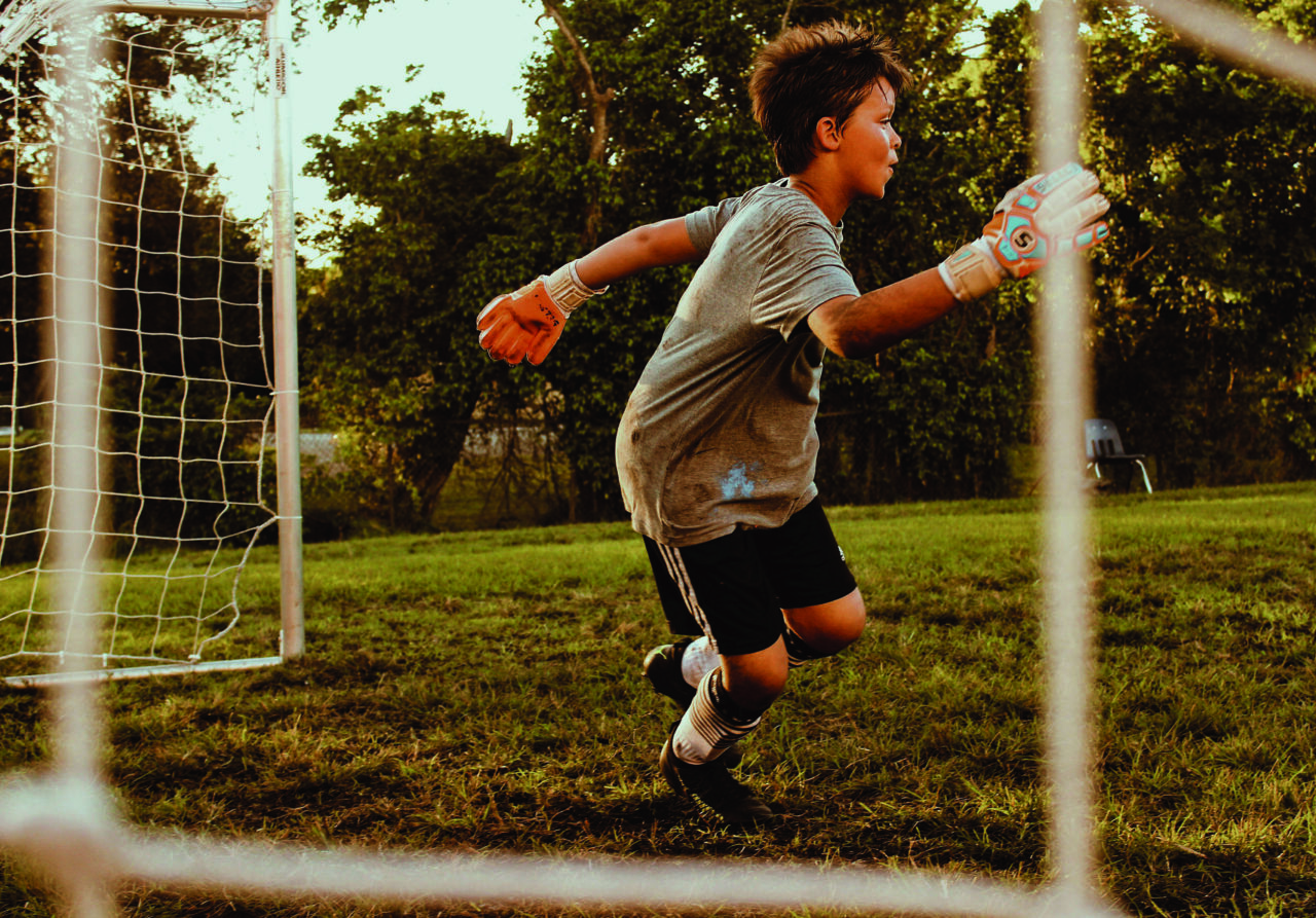 A young boy in a gray shirt and black shorts is running in a soccer field, wearing goalkeeping gloves. A soccer goal is visible in the background.