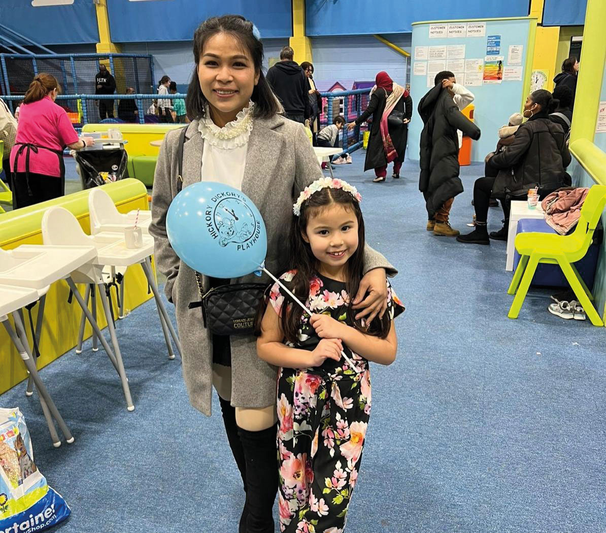 A woman and a girl stand together in a colorful indoor play area. The girl holds a blue balloon and smiles, while the woman wears a gray coat.