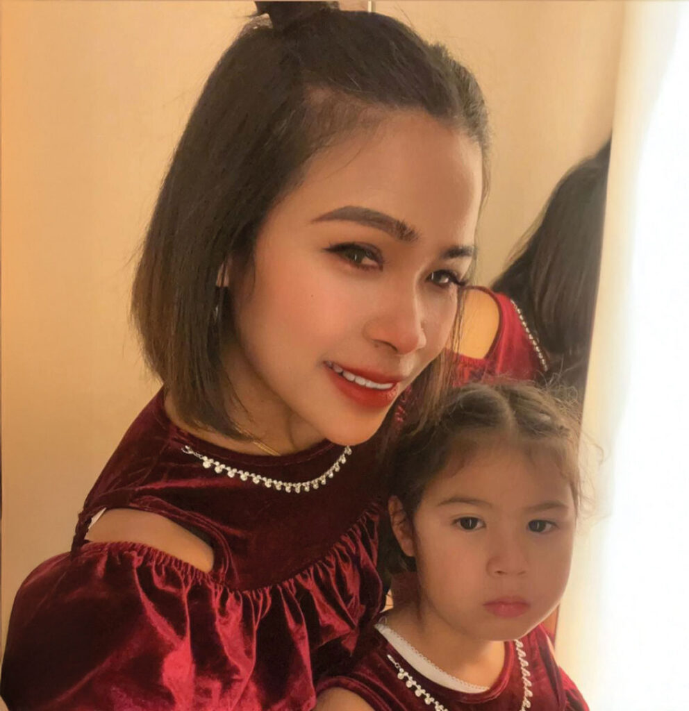 A woman and a young girl pose together, both wearing matching burgundy outfits with decorative details. The background features soft lighting and reflections.
