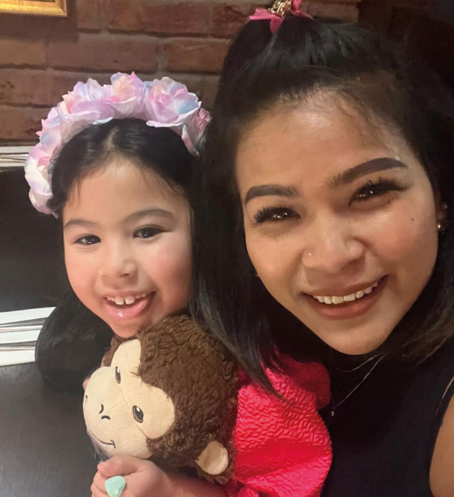A woman and a young girl smile together, with the girl holding a stuffed monkey toy. The girl wears a floral headband.