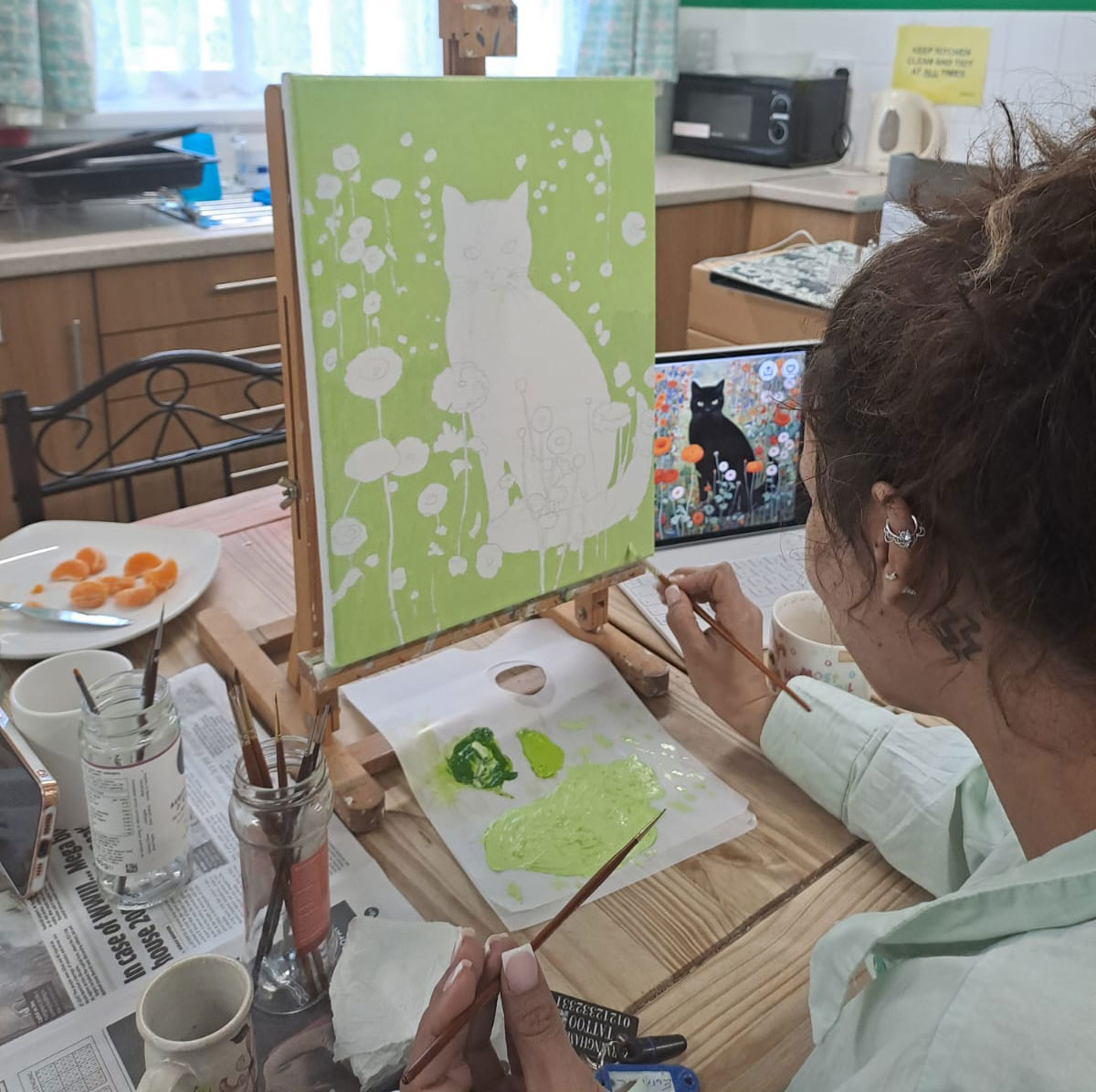 An artist is painting a green canvas featuring a white cat and flowers, with paintbrushes in hand. A reference image of a black cat is visible on a nearby screen.