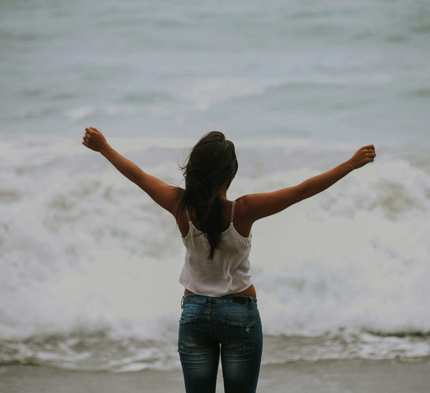 A person stands on the beach with arms raised, facing the ocean waves, wearing a white top and blue jeans.