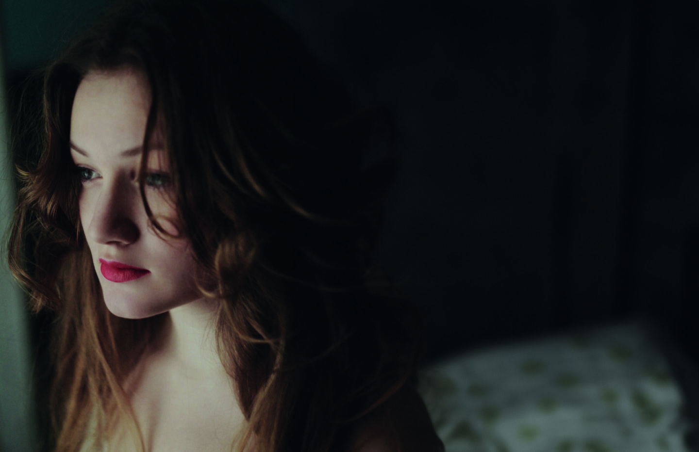 A young woman with wavy hair gazes thoughtfully to the side, her lips painted red, against a dark background.