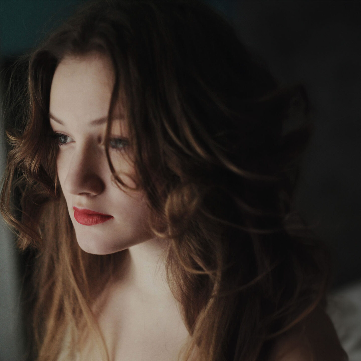 A young woman with long, wavy hair and red lipstick gazes thoughtfully to the side, with soft lighting highlighting her features.