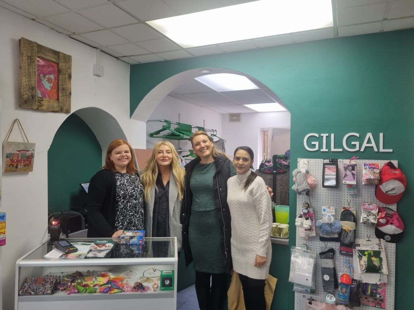 Four women stand together in a shop with a green wall. They are smiling, with a display of various items and the word "GILGAL" visible on the wall.