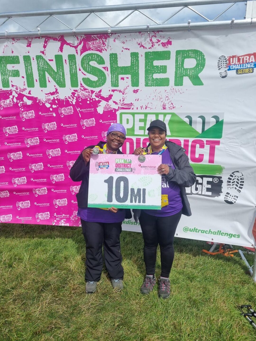 Two women stand together, smiling and holding a sign that reads "10 MI," in front of a colorful "FINISHER" banner at an event. They are wearing matching purple shirts and outdoor gear.