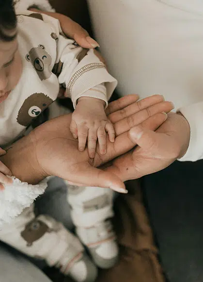 Close-up of an adult gently holding a baby's hand. The baby is wearing a cream-colored outfit with bear illustrations.
