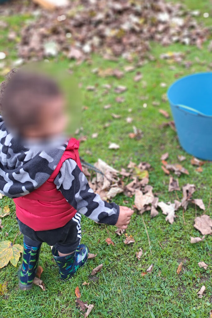 A child in a red vest and camouflage hoodie stands on grass, holding a leaf, with a blue bucket nearby and a pile of leaves in the background.