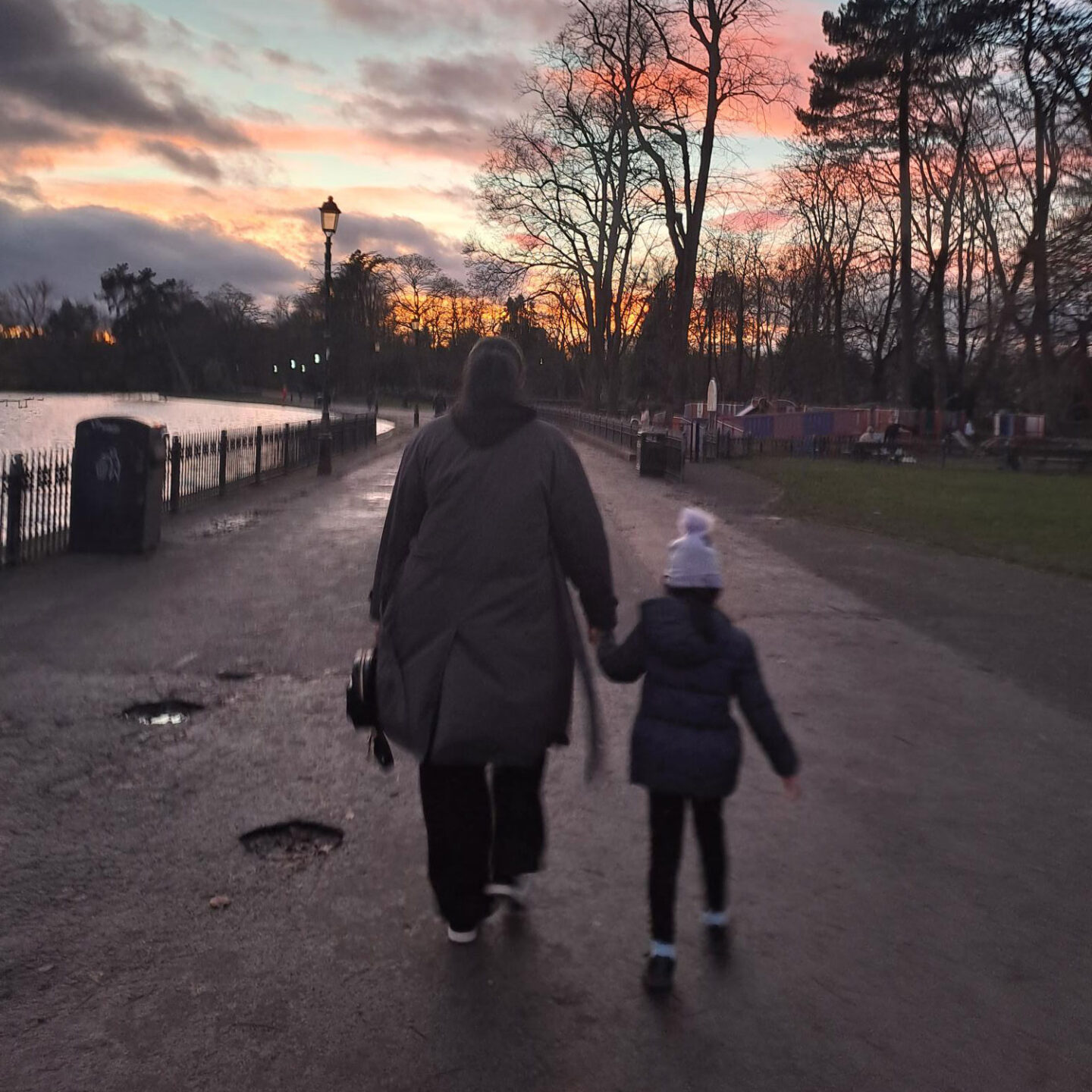 A person and a child walk hand in hand along a path by a lake at sunset, with trees and a park in the background.