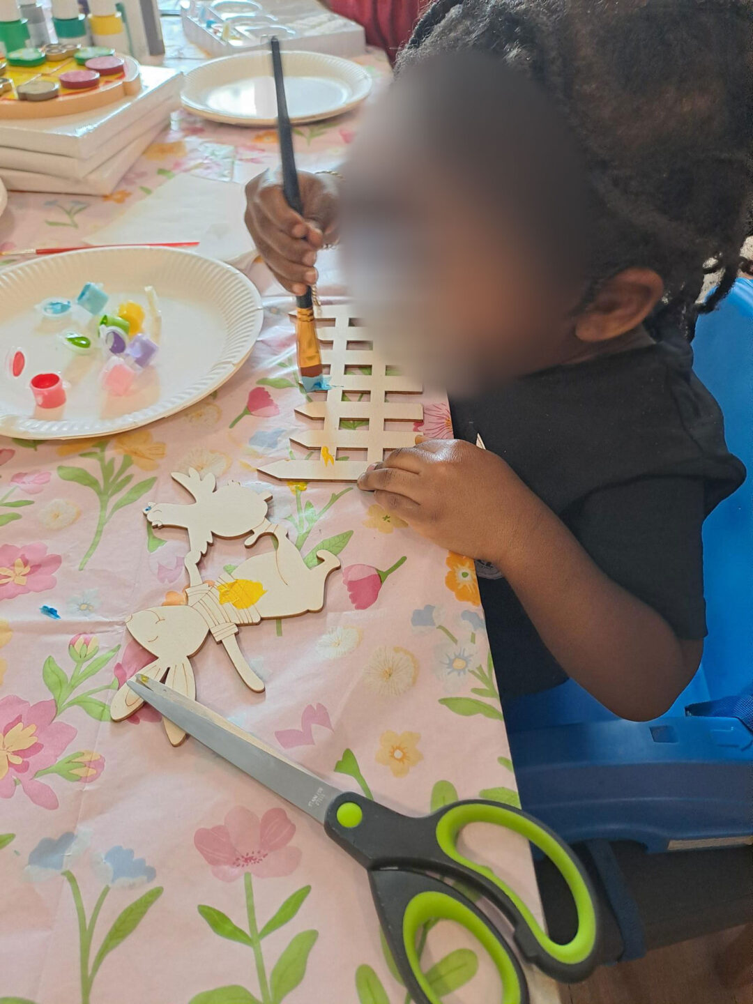 A child is painting a wooden craft piece at a table covered with floral-patterned paper. Scissors and colorful paint containers are nearby.