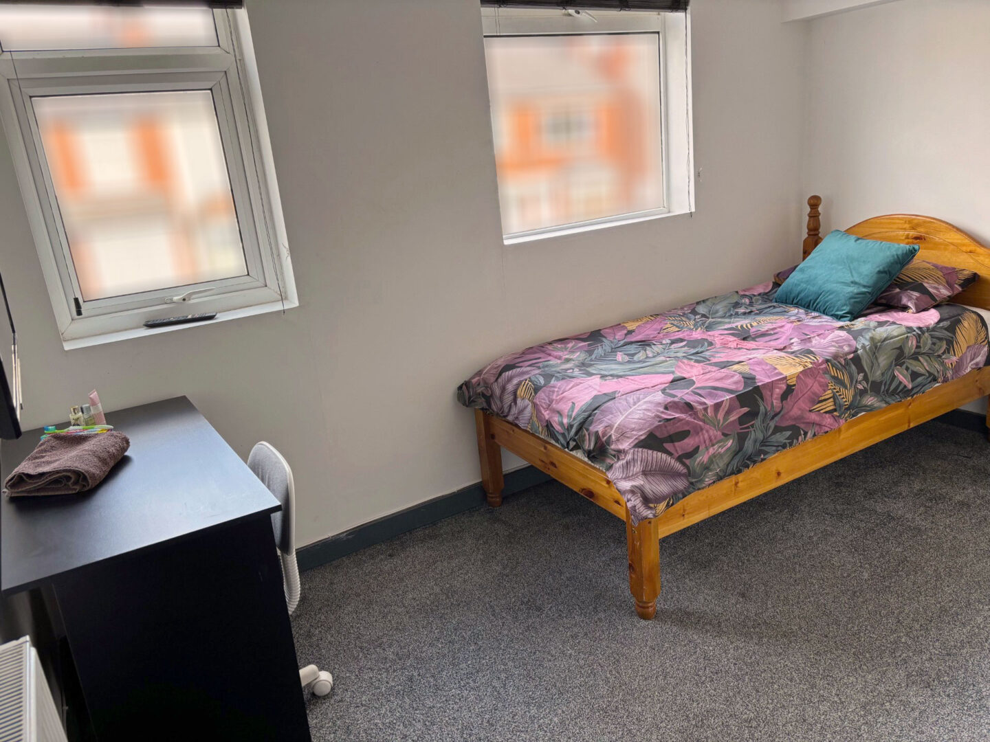 A cosy bedroom featuring a single bed with a floral-patterned duvet, a teal pillow, and a desk with a chair. Two windows allow natural light to enter the room.