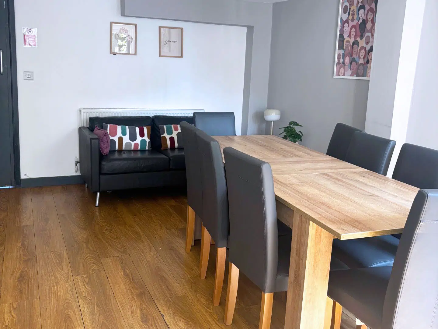 A modern dining area featuring a wooden table surrounded by black leather chairs. A black sofa with colorful cushions is in the background, along with framed artwork on the walls.