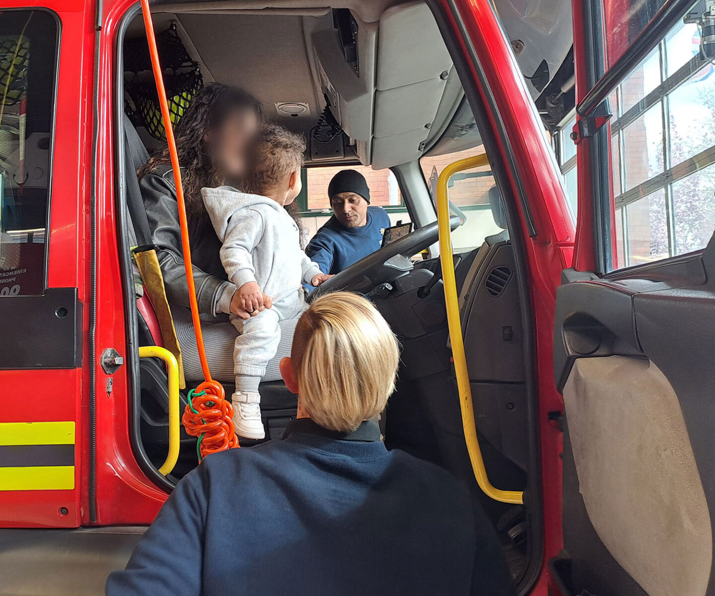A child sits on the lap of an adult in the driver's seat of a fire engine, while another adult engages with them.