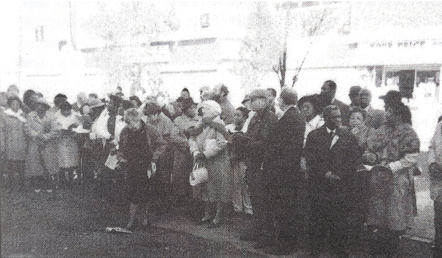A crowd of people, mostly dressed in coats and hats, stand in the street celebrating.