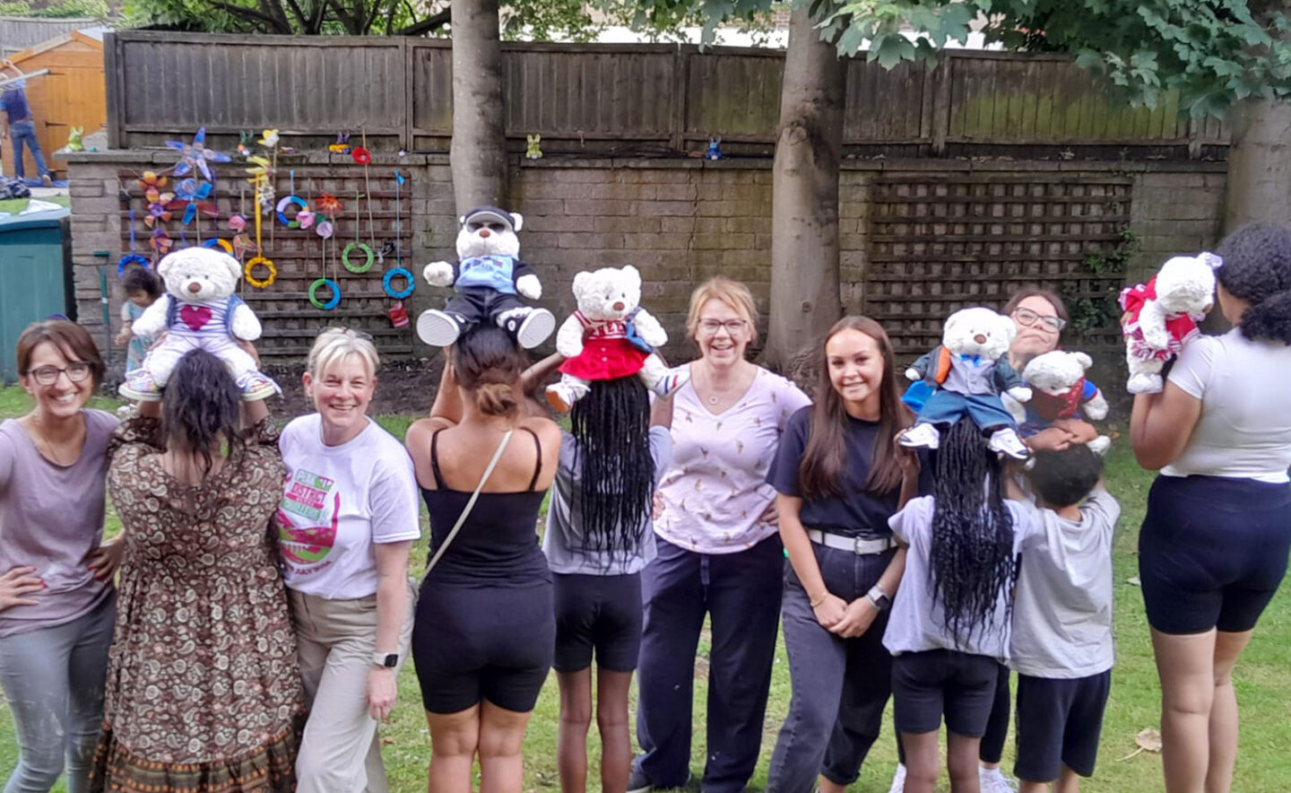 A group of women and children stand outdoors, holding teddy bears on their heads.