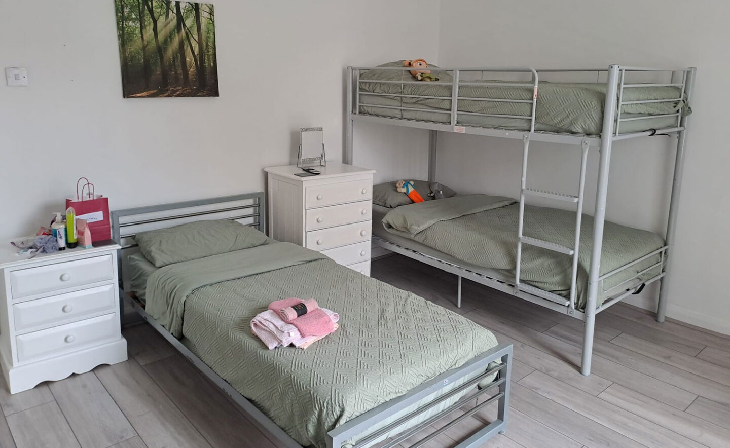 A clean and welcoming bedroom featuring a single bed and a set of bunk beds, all with sage green bedding. A white dresser and bedside table are present, along with some toys and personal items. A nature-themed wall art is displayed above the beds.