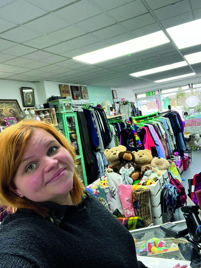 A woman with shoulder-length reddish hair smiles in a thrift store filled with clothing racks and stuffed animals.