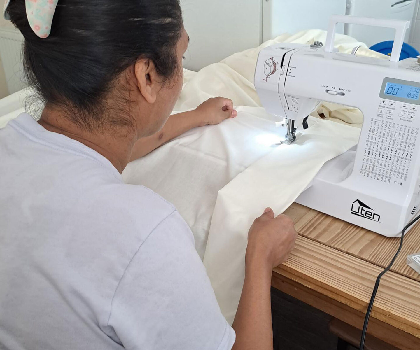 A woman is sewing white fabric using a sewing machine on a wooden table.