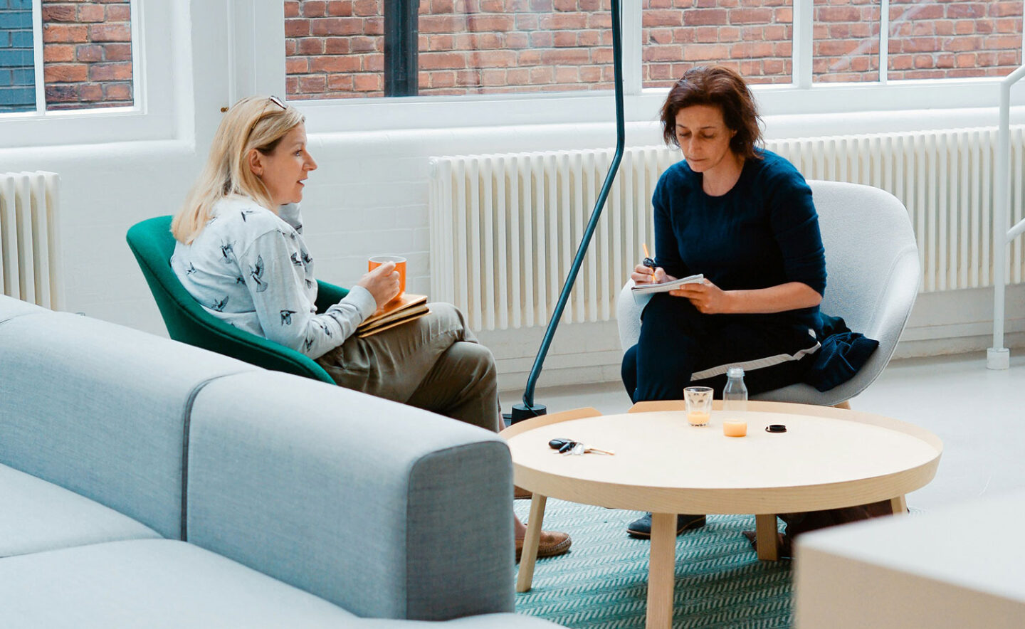 Two women are seated in a modern, bright room, engaged in conversation. One woman holds a cup and notebook, while the other writes notes. A round coffee table with a few items is in front of them.