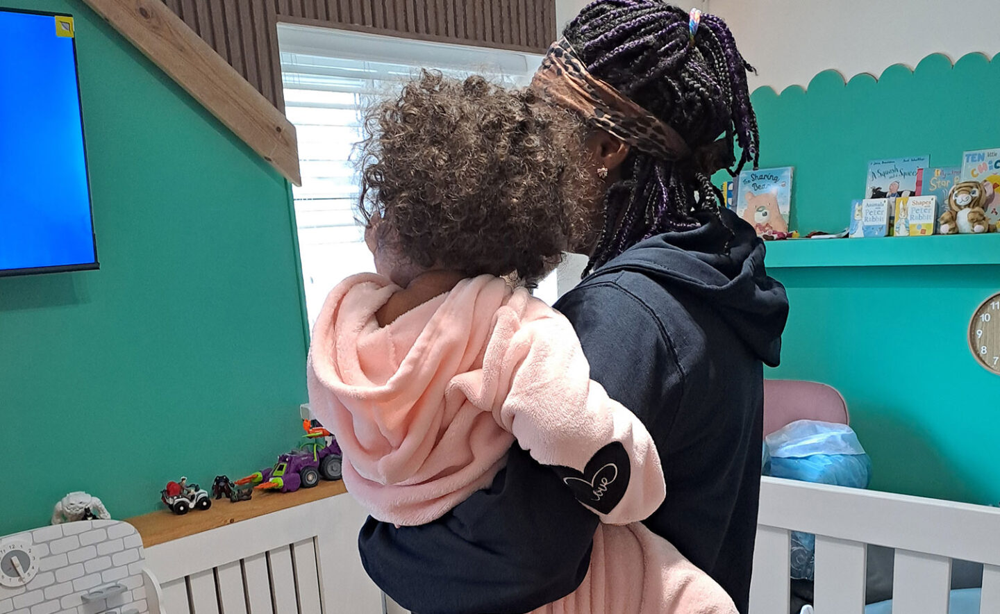 A woman holds a child in a cosy room with green walls, toys on a shelf, and a television in the background. The child is wearing a pink hoodie.