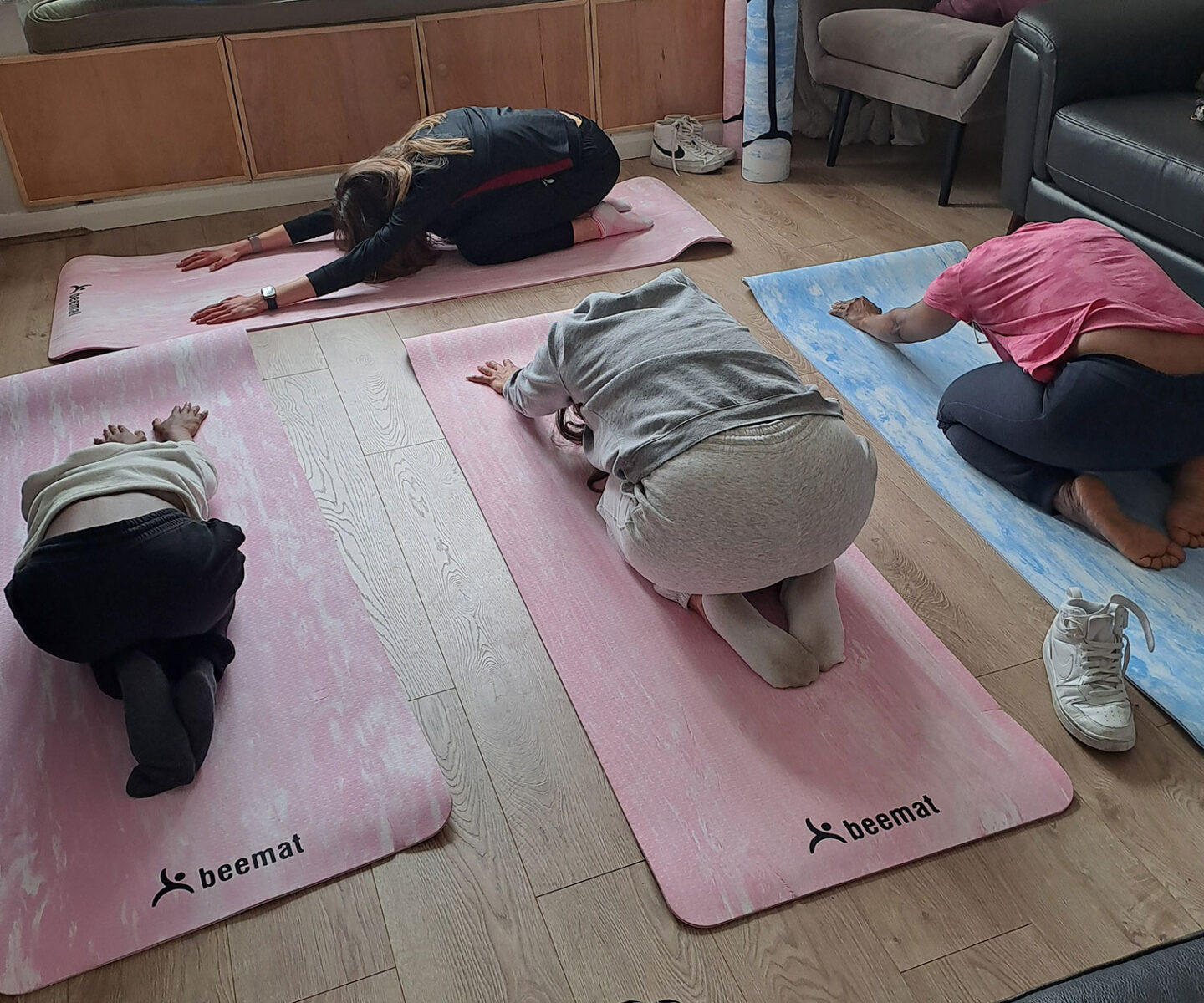 Three women are practicing yoga, led by a fourth woman. They each in a child's pose position, on mats in shades of pink and blue.
