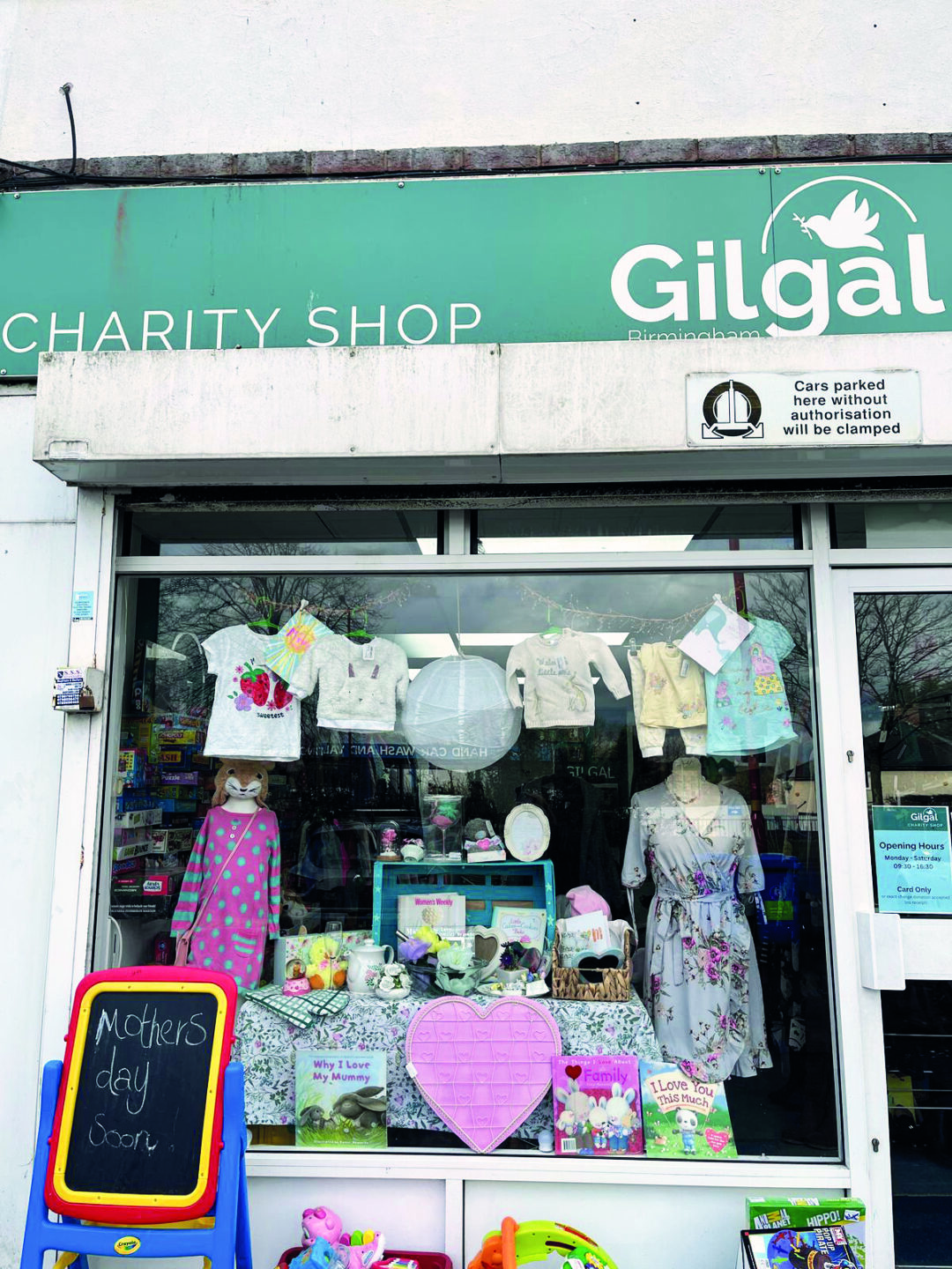 A charity shop window display featuring children's clothing, toys, and Mother's Day decorations. A chalkboard sign reads "Mother's Day Soon.