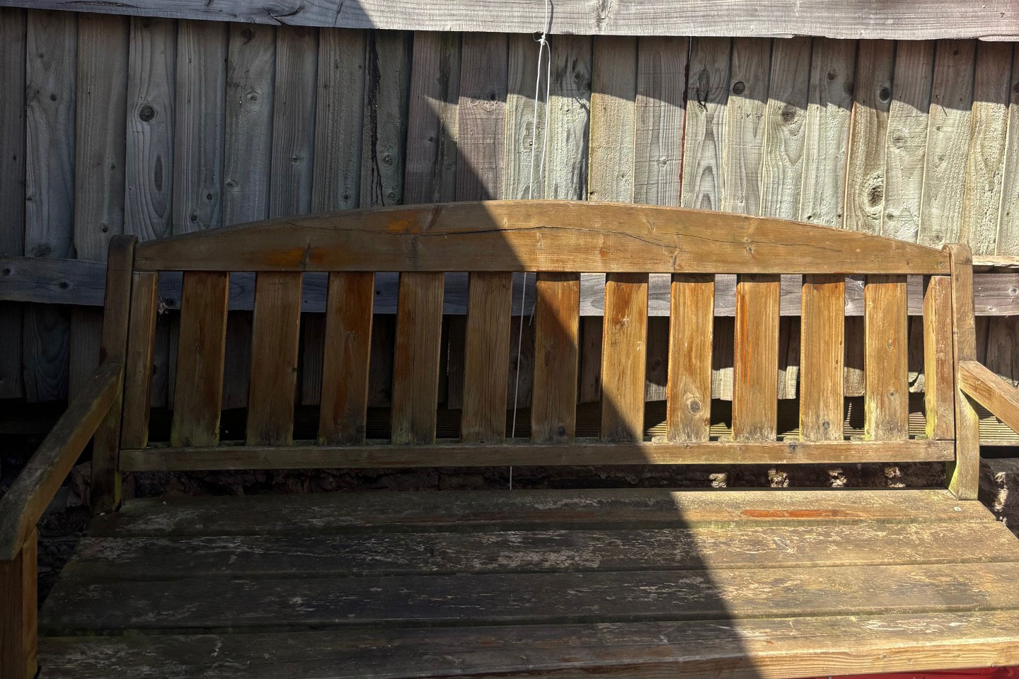 A weathered wooden bench with slatted backrest and seat, positioned against a wooden fence. Sunlight casts a shadow on the bench.