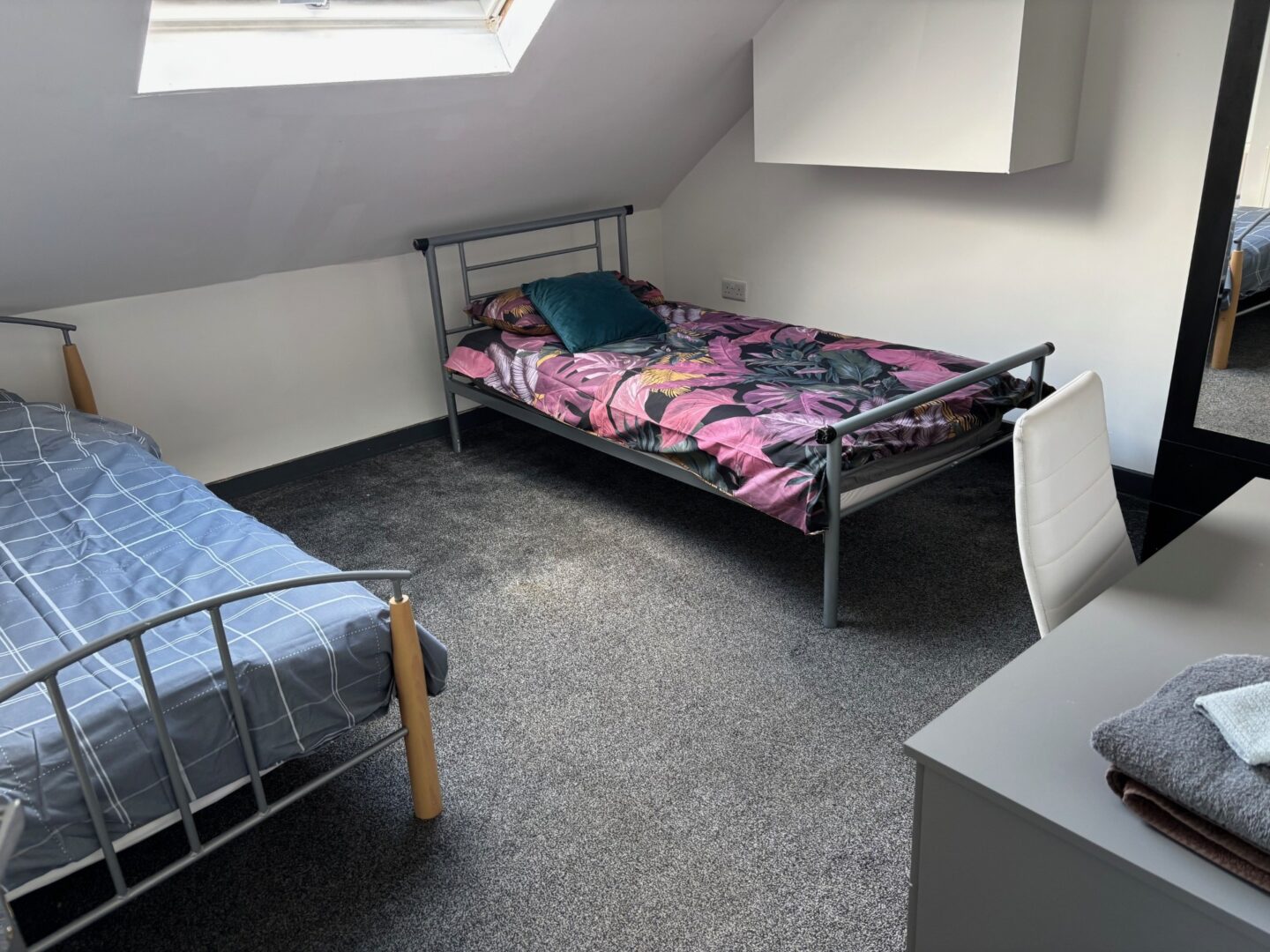 A clean and tidy bedroom featuring two metal-framed single beds, a desk, chair, and mirror. A skylight provides natural light.