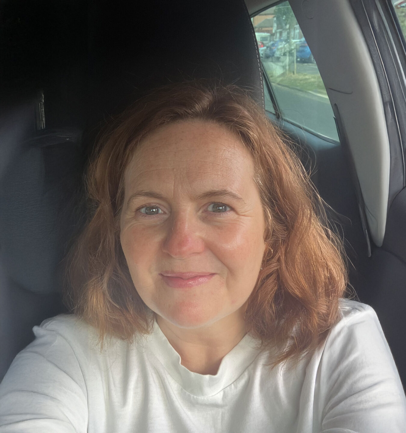 A woman with shoulder-length, wavy brown hair smiles at the camera while sitting in a car. Natural light illuminates her face.