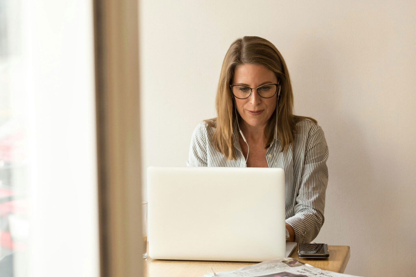 A woman with long hair and glasses is focused on her laptop while wearing earphones, sitting at a wooden table. A glass of water and a smartphone are nearby.