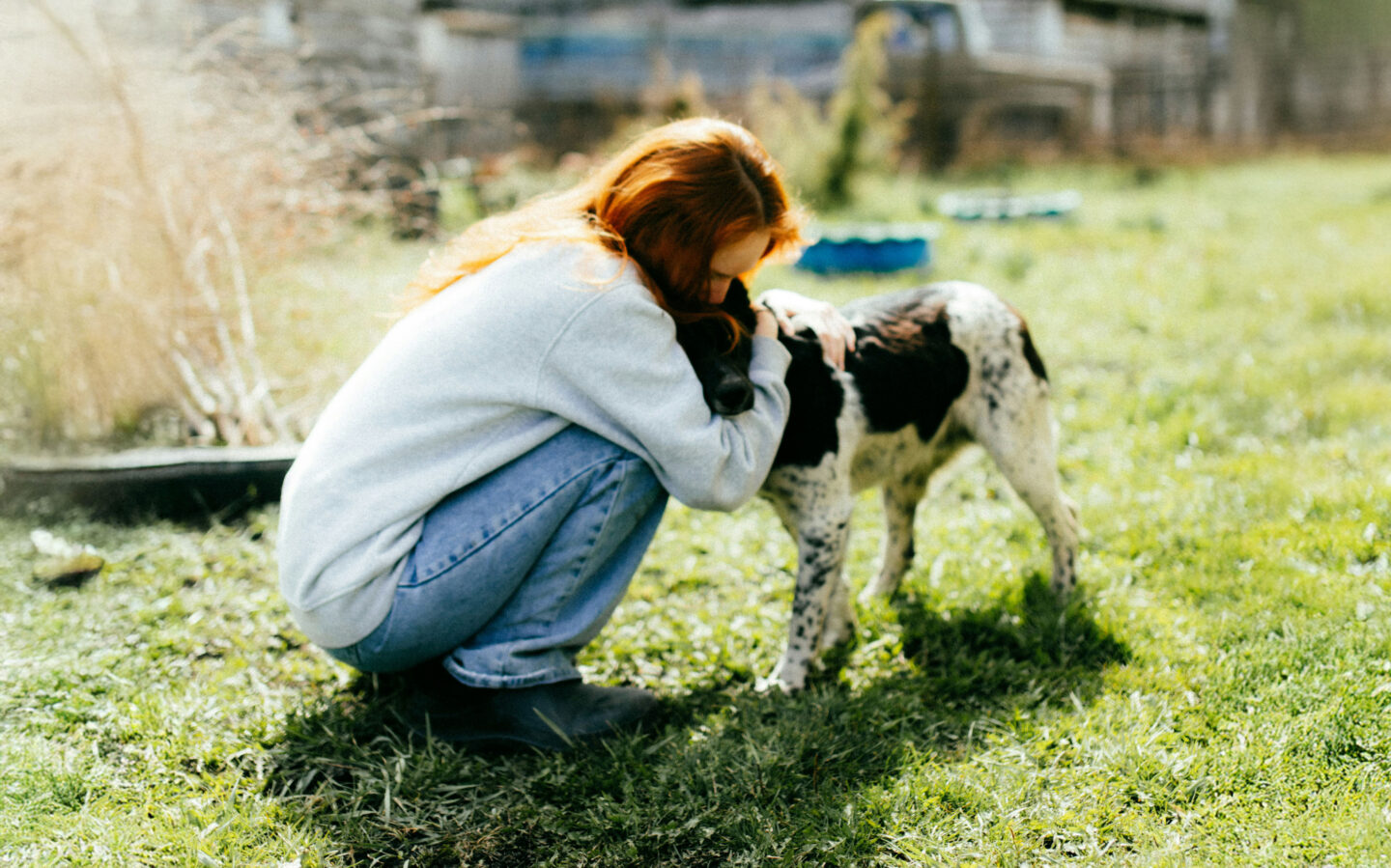 A person with long red hair kneels on grass, hugging a black and white dog. The background features greenery and a blurred structure.