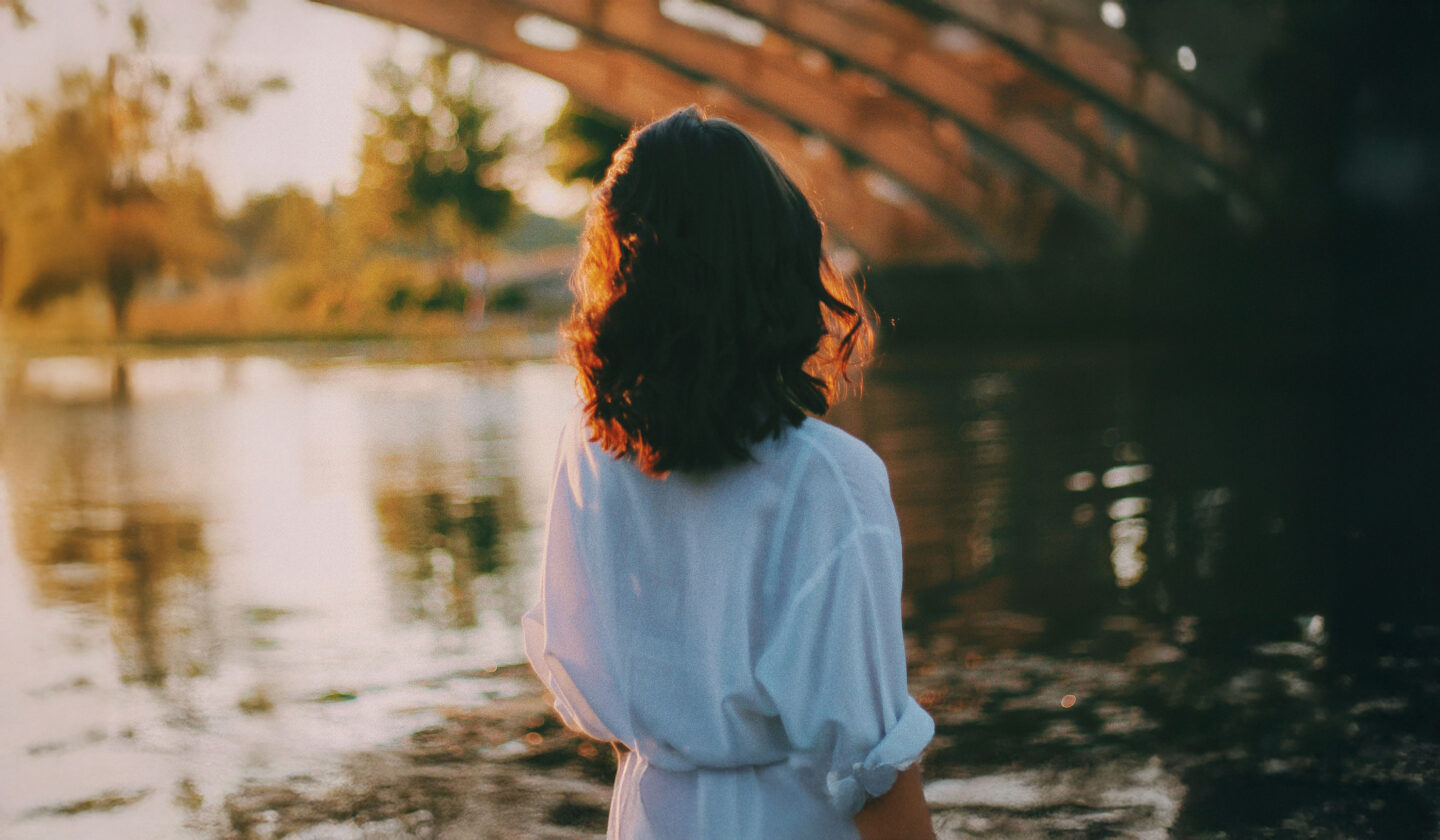 A person with curly hair stands by a calm body of water, facing away, with sunlight illuminating their hair and a bridge in the background.