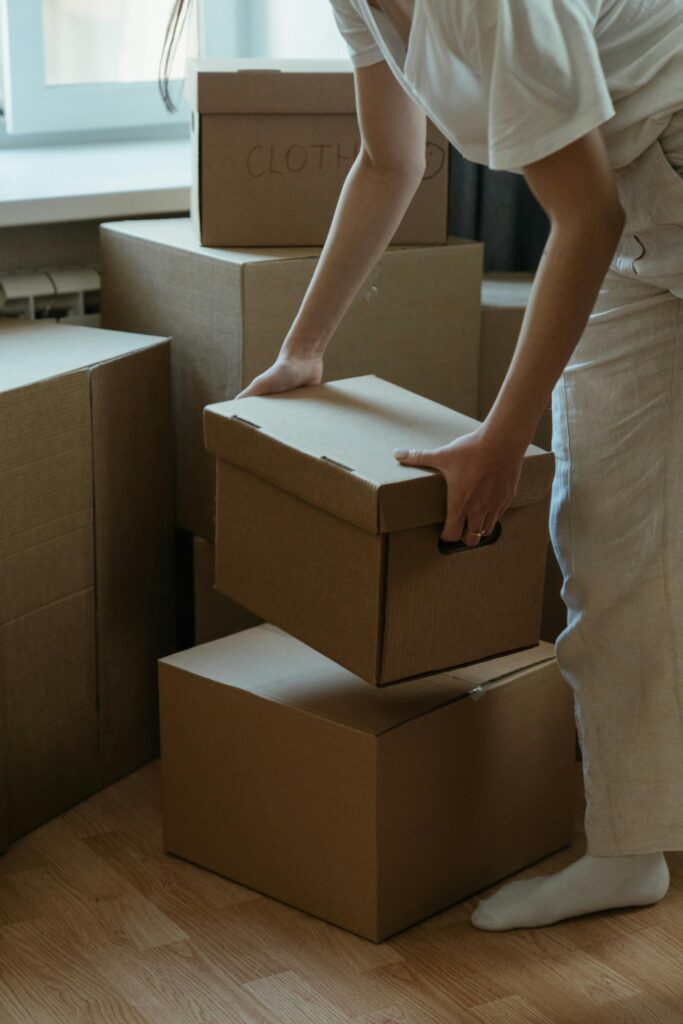A person is lifting a cardboard box while surrounded by several stacked boxes in a room with natural light.