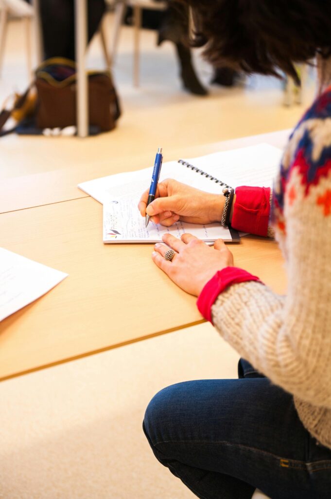 A person is writing in a notebook with a blue pen, seated at a table. They are wearing a cozy sweater and a red shirt underneath.