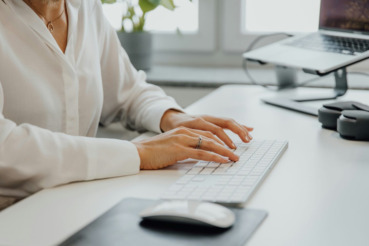 A person typing on a keyboard with a computer mouse and headphones on a desk, surrounded by plants and natural light.