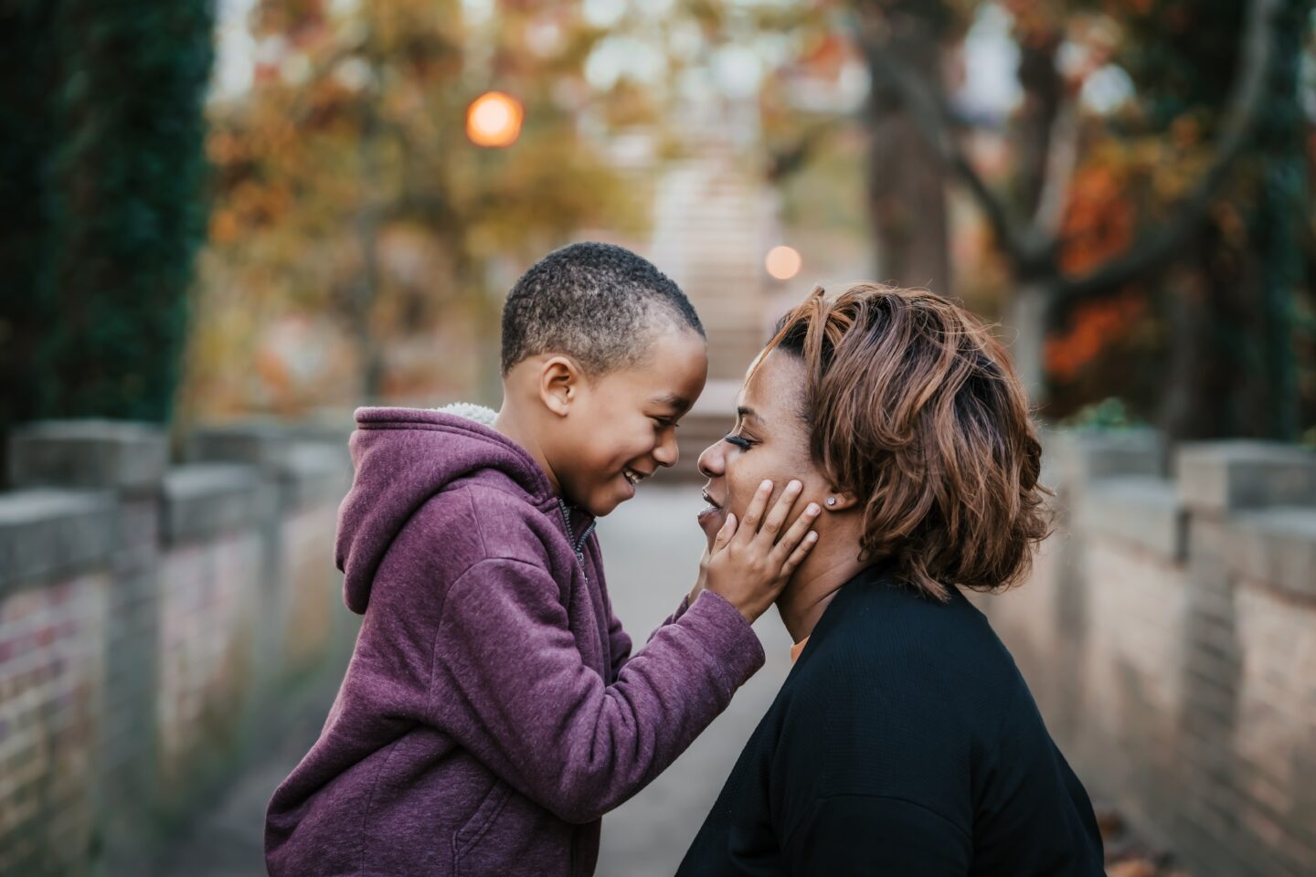 A child and an adult woman share a joyful moment, smiling and touching each other's faces in a park setting with autumn foliage.