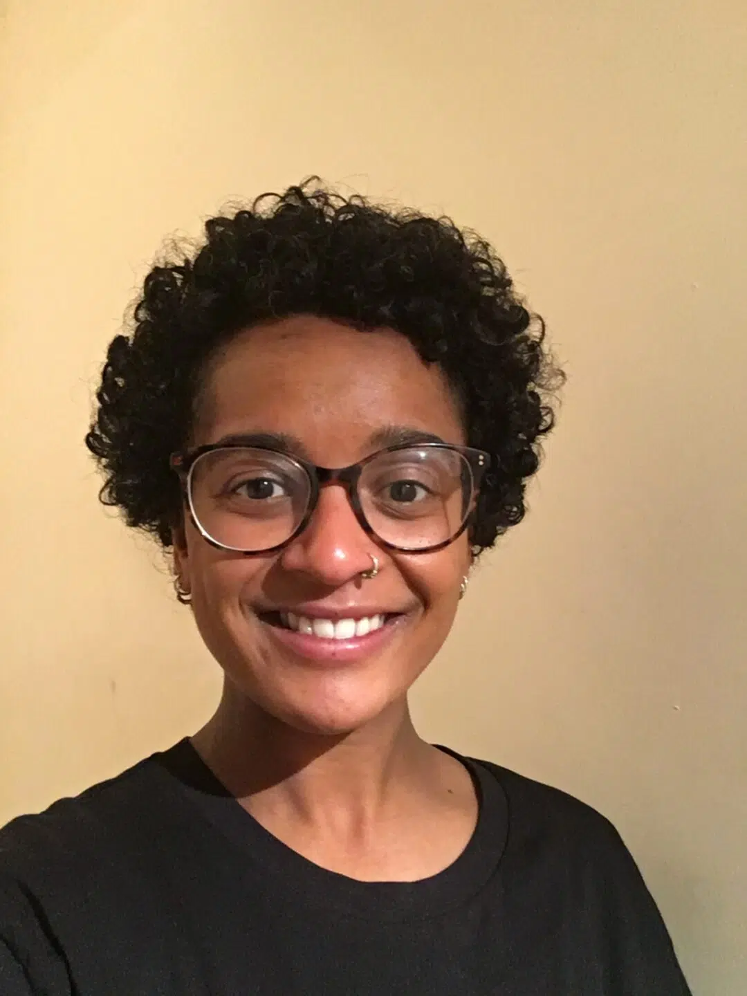 A person with short, curly hair and glasses smiles at the camera, wearing a black shirt against a plain background.
