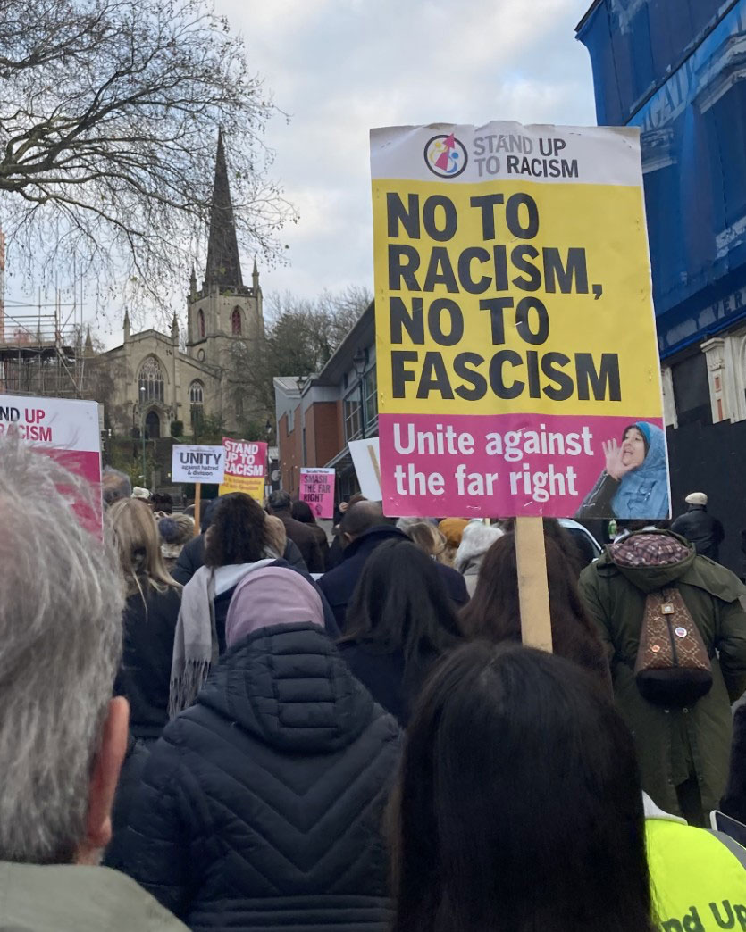 A crowd of people holding signs at a protest, with a prominent sign reading "No to Racism, No to Fascism" and "Unite against the far right." A church is visible in the background.
