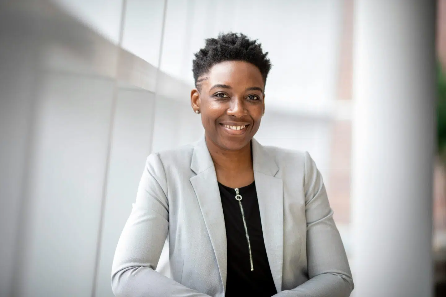 A smiling woman with short, curly hair is seated, wearing a light gray blazer over a black top, against a bright, modern background.