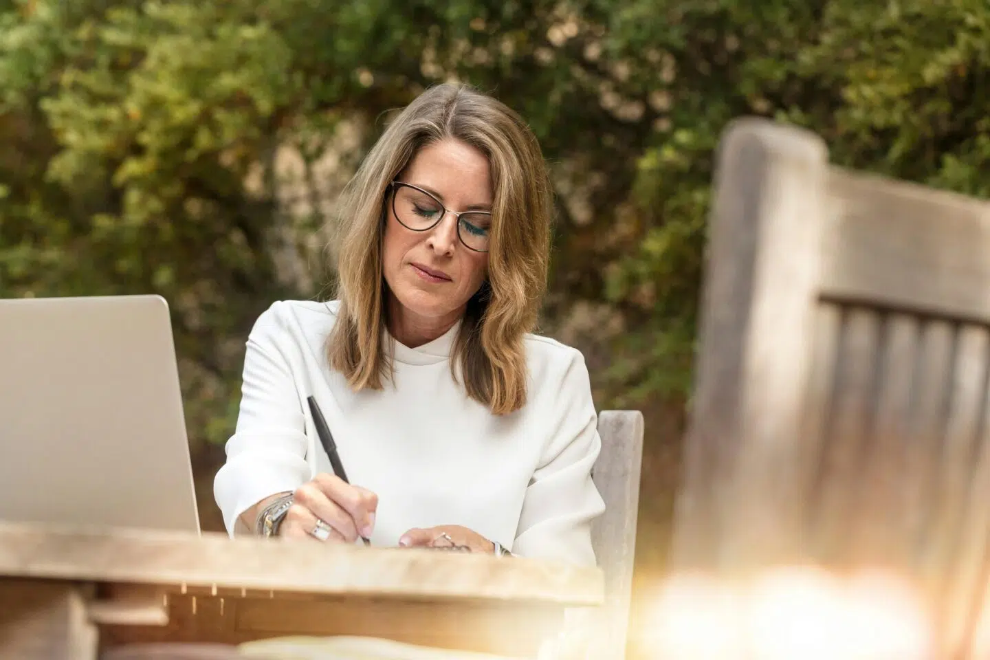 A woman with glasses is sitting at a wooden table outdoors, writing with a pen. A laptop is open beside her, and greenery is visible in the background.