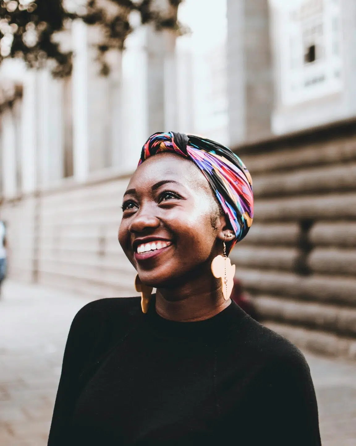A smiling woman wearing a colorful headscarf and large earrings stands outdoors, with a blurred background of a stone wall.