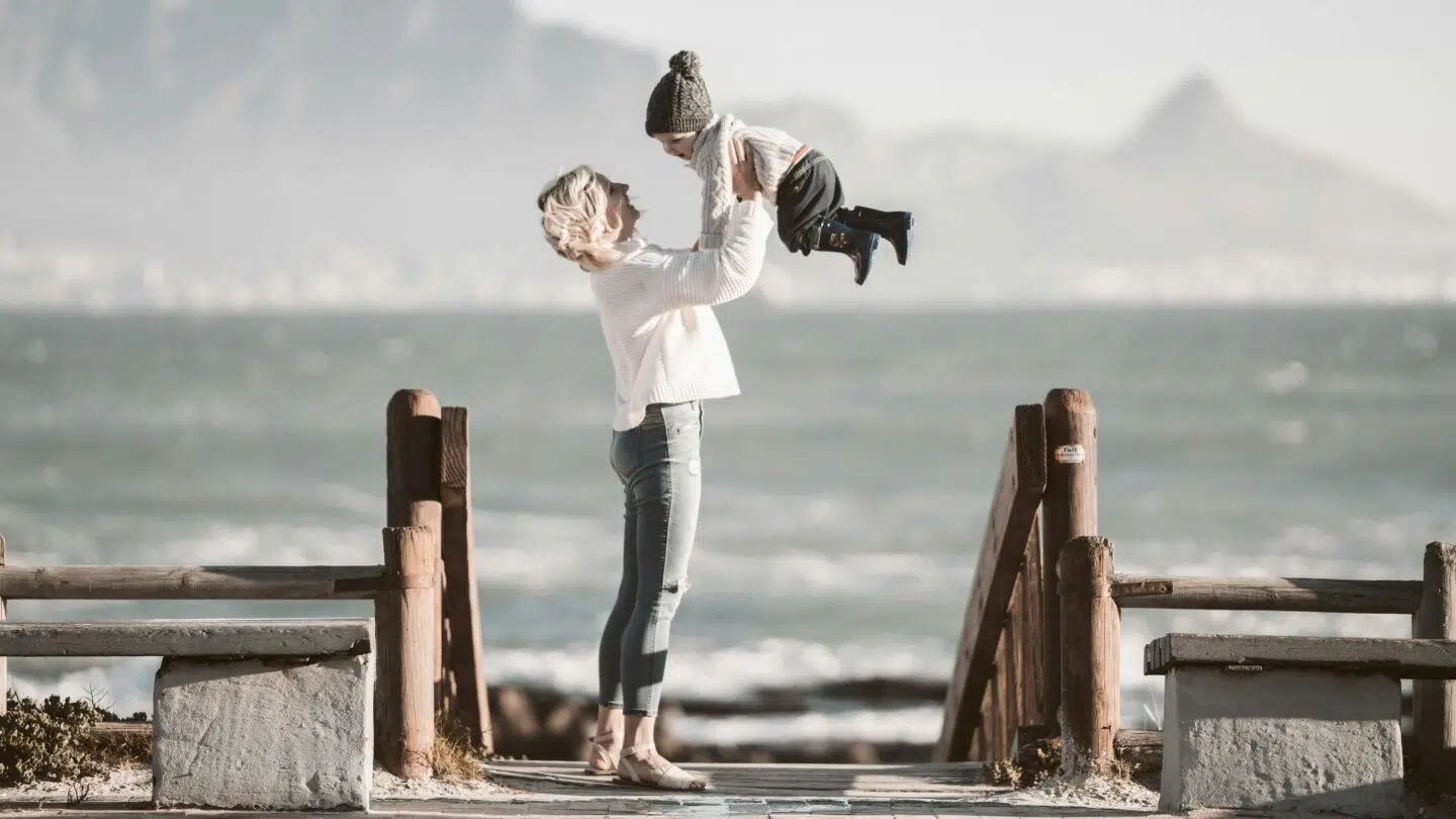 A woman lifts a child joyfully on a wooden walkway by the beach, with waves and mountains in the background.