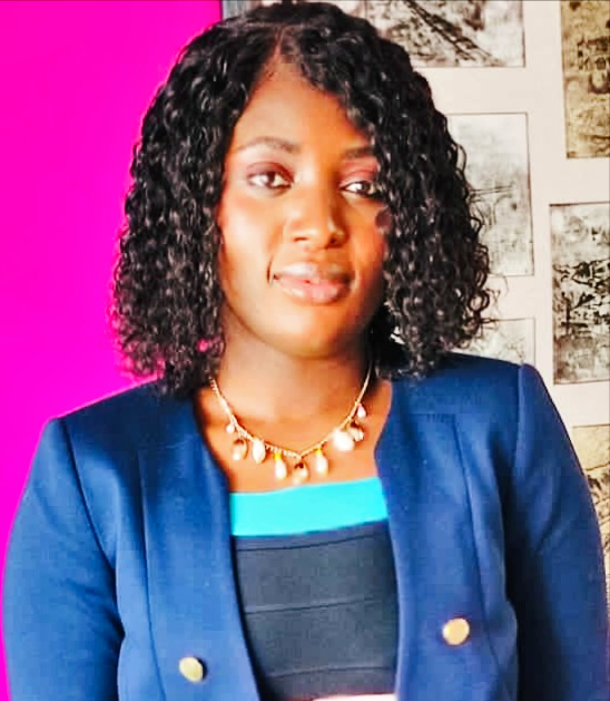 A woman with curly hair is wearing a blue blazer and a necklace, standing in front of a pink wall.